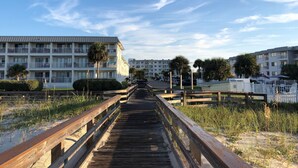 A wooden boardwalk leading to a beachfront hotel with multiple balconies and palm trees.