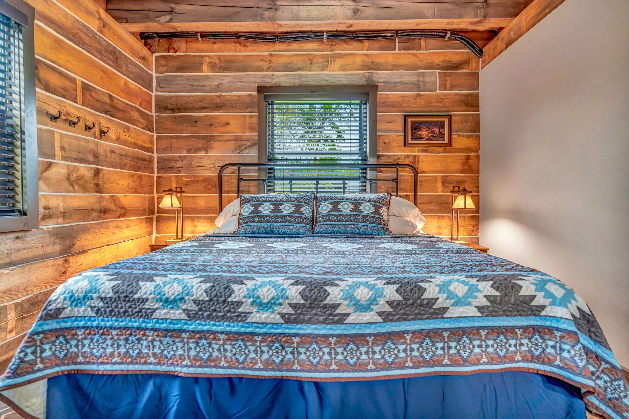 A wooden cabin room with a bed, two wall-mounted lamps, and a window with blinds.