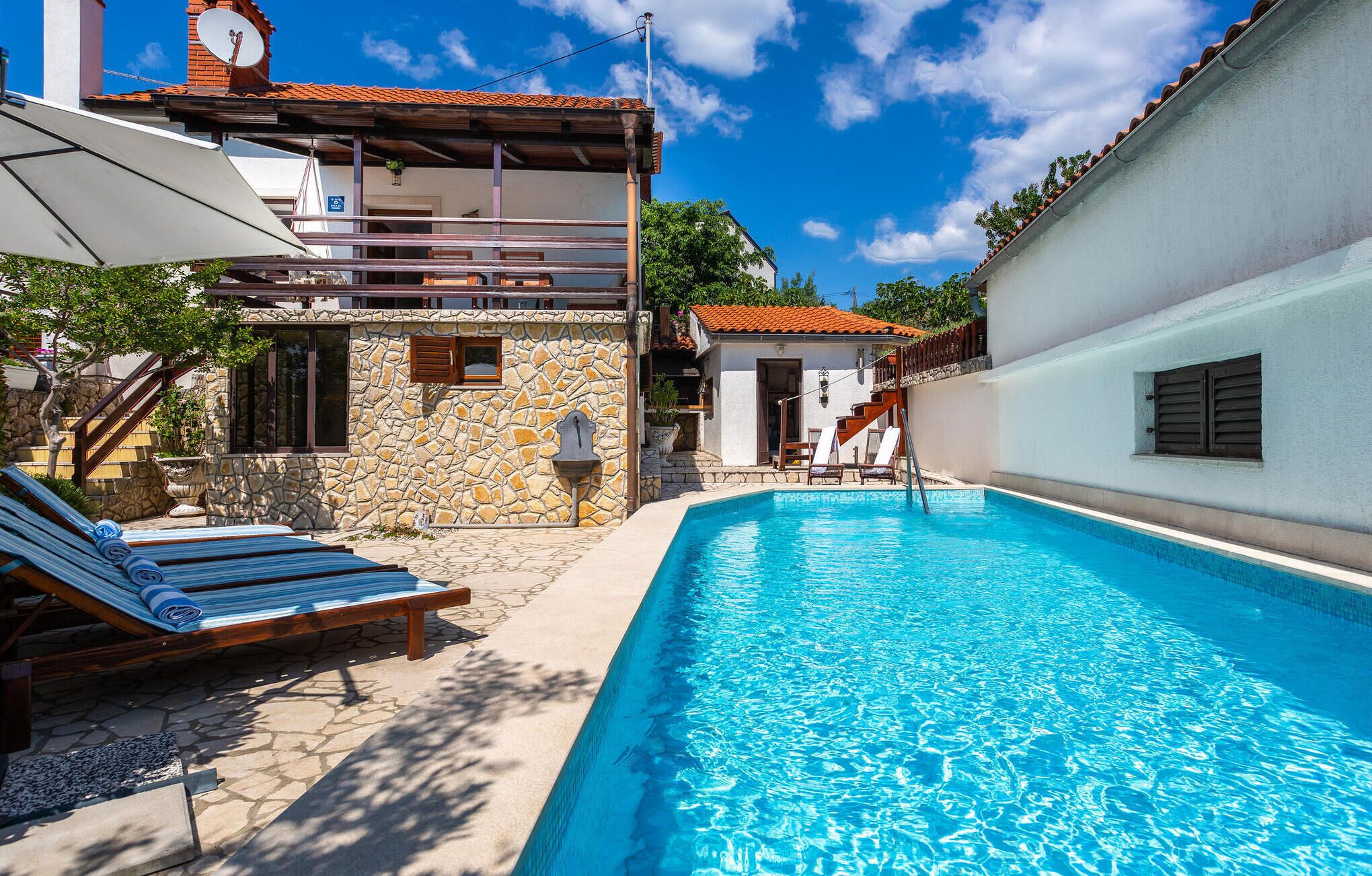 A swimming pool with lounge chairs, a stone building, and a white wall with shutters.