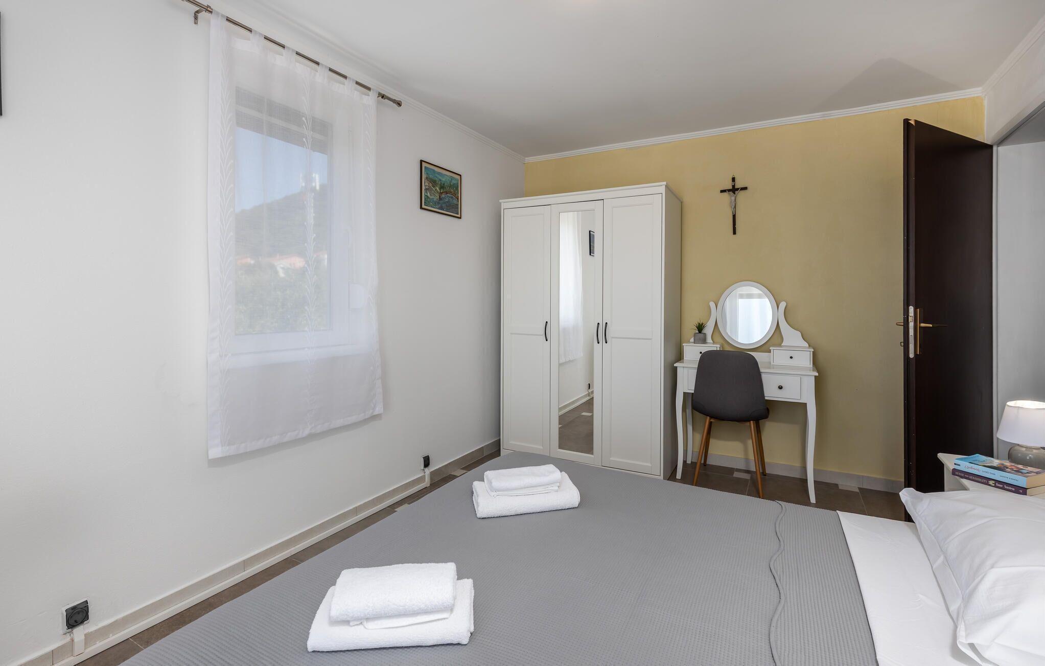 A hotel room with a bed, a desk with a mirror, a wardrobe, and a cross on the wall.