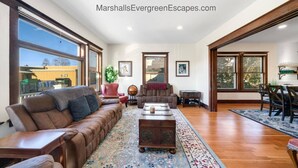 A spacious living room with a brown leather sofa, a wooden coffee table, and a dining area with a table and chairs. Large windows provide ample natural light and views of the outside. - Mountain Manor in Sandy| HotTub |3KingBeds| MtHood (Sandy)