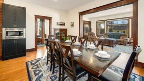 A dining area with a wooden table set for four, a kitchen with a built-in microwave, and a view into a living room with a sofa and chairs. - Mountain Manor in Sandy| HotTub |3KingBeds| MtHood (Sandy)