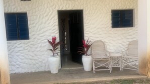 A white building with textured walls, a doorway, and two potted plants with red leaves. There are two white wicker chairs outside. - Beach-side one bedroom (Kwale County)