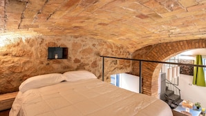 Un dormitorio con techo de ladrillo, una cama con ropa blanca, un estante pequeño con un recipiente, y vistas de una escalera y una cortina verde.