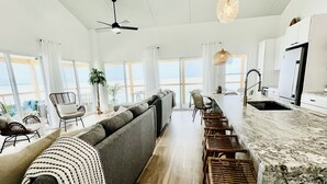 Een moderne woonkamer met een grijze bank, houten vloeren en een keuken met witte kasten en een marmeren aanrecht.