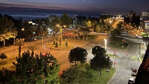 Vista aérea de uma paisagem urbana noturna com ruas iluminadas, uma rotatória e um corpo d'água ao longe.
