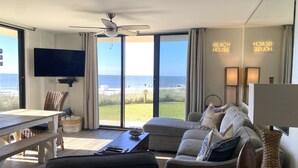 A modern living room with a large window overlooking the beach, a ceiling fan, and a flat-screen TV. - Beautiful Ground Floor with a Panoramic View of the Gulf of Mexico (Orange Beach)