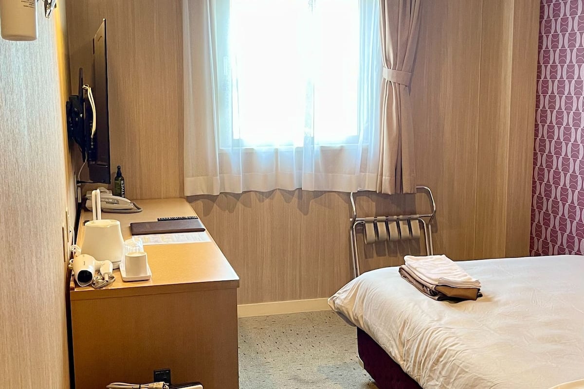 A hotel room with a bed, a desk with a computer, a chair, and a window with curtains.