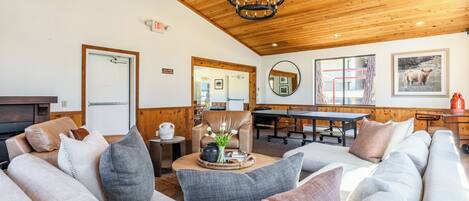 A cozy living room with a wooden ceiling, a round mirror, a billiards table, and a sofa with cushions.