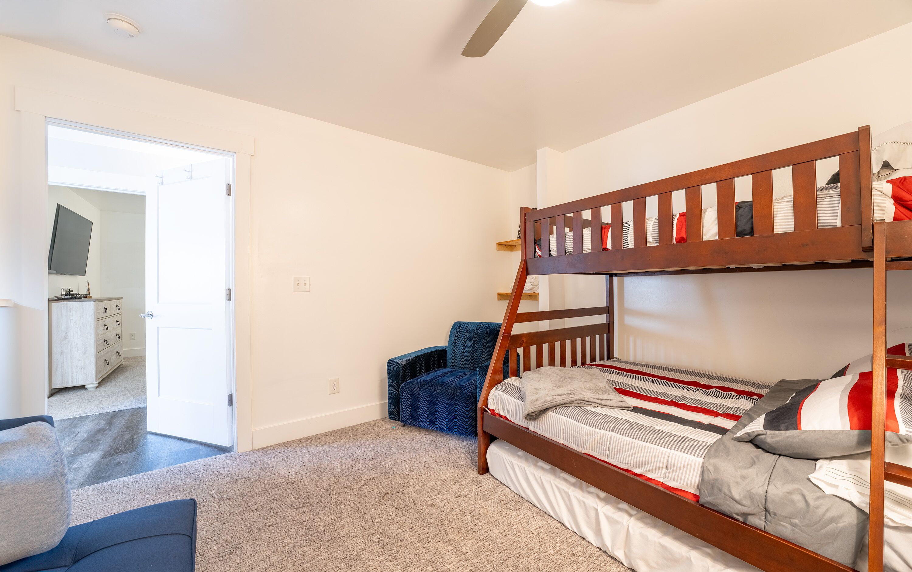 A bunk bed with a wooden frame, a blue chair, and a white dresser in a room with white walls and carpeted floor.