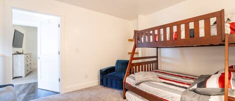 A bunk bed with a wooden frame, a blue chair, and a white dresser in a room with white walls and carpeted floor.