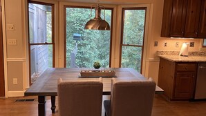 Une cuisine avec une table en bois, deux chaises, un luminaire suspendu et de grandes fenêtres donnant sur un paysage verdoyant.