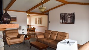 Ein Wohnzimmer mit einem braunen Ledersofa, einem braunen Ledersessel und einem Couchtisch aus Holz. Auf einem Holzregal befinden sich eine weiße Lampe und eine dekorative Vase.
