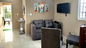 Una sala de estar con un sofá gris, una mesita blanca y un televisor montado en la pared.
