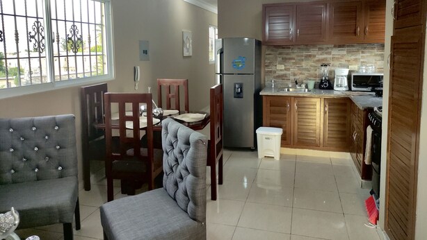 Una cocina compacta con armarios de madera, un refrigerador, un microondas y una cafetera. Hay una zona de comedor con dos sillas y una mesa.