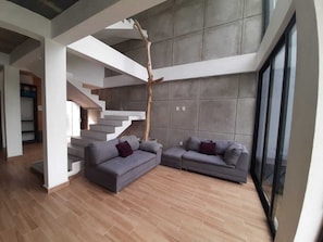 A modern living room with a staircase, grey sofas, and a large window.