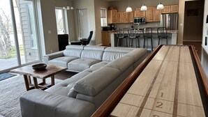 A modern living room with a large sectional sofa, a shuffleboard table, and a kitchen area with wooden cabinets and stainless steel appliances. - Stillwater Riverfront Home, Close to the Beartooths, Fish from your door - Peace (Absarokee)