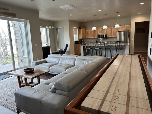 A modern living room with a large sectional sofa, a shuffleboard table, and a kitchen area with wooden cabinets and stainless steel appliances. - Stillwater Riverfront Home, Close to the Beartooths, Fish from your door - Peace (Absarokee)