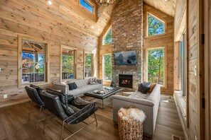 Cozy living area with a high ceiling, fireplace, and large windows offering views of the surrounding landscape.