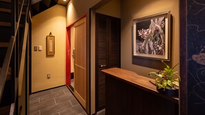 A hallway with a wooden staircase, a red door, a wall-mounted picture, and a wooden counter with a plant. - TSUDOYA Sannou (Osaka)