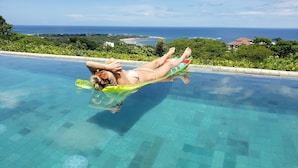 Una persona recostada en un flotador en una piscina cristalina, con vista al océano y follaje en el fondo.