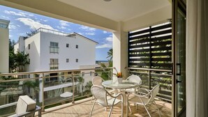 Un balcon doté d’une balustrade en verre, une table ronde avec des chaises, et une vue sur un bâtiment blanc.