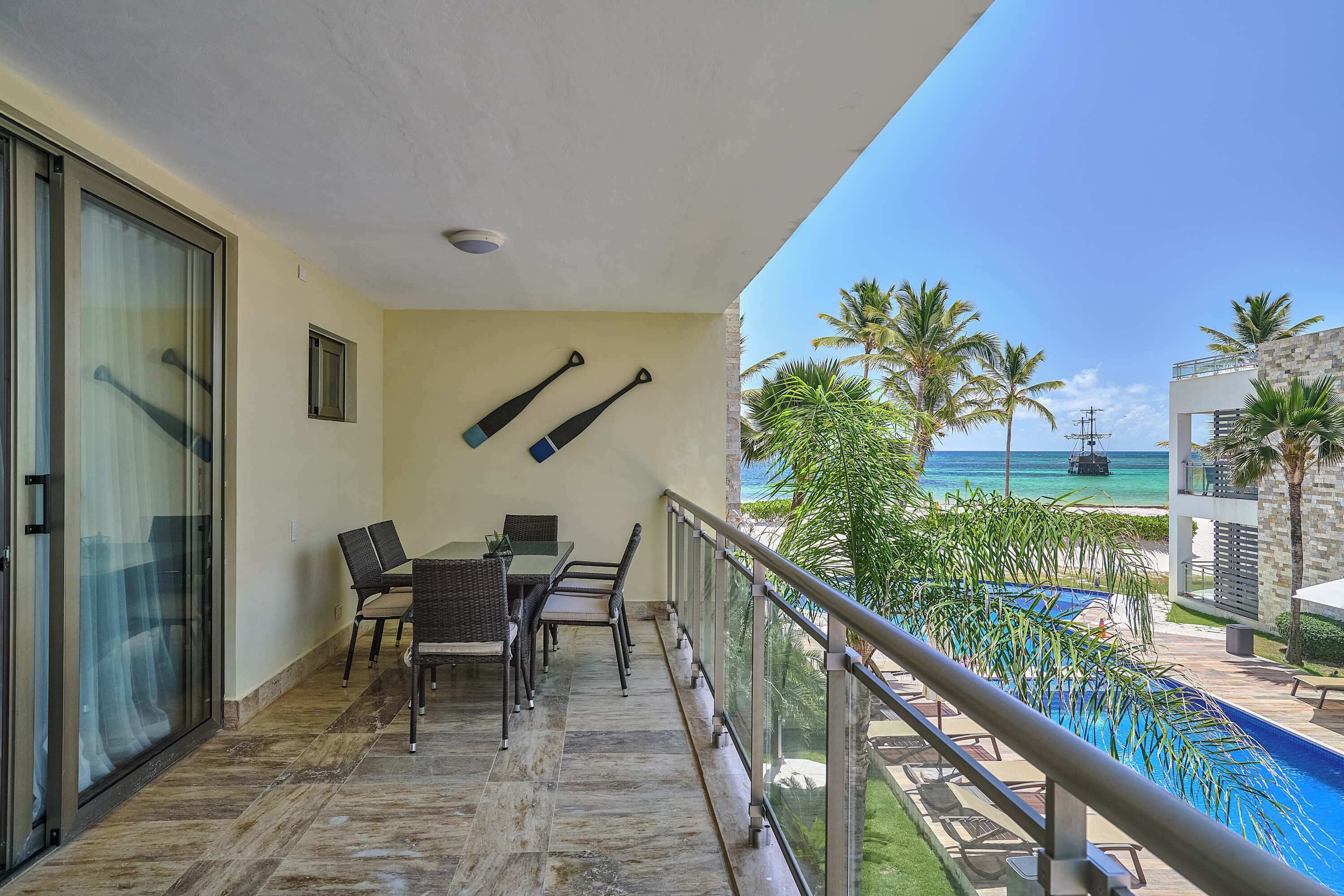 Uma varanda com mesa e cadeiras, uma porta de vidro e vista para a piscina e a praia.