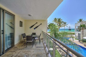 Un balcón con mesa y sillas, un vidrio y vista a la piscina y la playa.