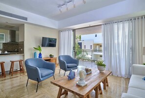 Ein modernes Wohnzimmer mit Couchtisch aus Holz, zwei blaue Sessel, einem weißen Sofa, einem an der Wand montierten Fernseher und einem Balkon mit weißen Vorhängen.