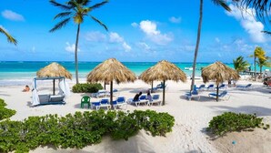 Ein Strand mit reetgedeckten Sonnenschirmen, Liegestühlen und Palmen.