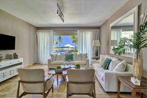 A living room with a sofa, chairs, a coffee table, a TV, and a view of palm trees and a pool.