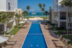 Una piscina con sillas de descanso y sombrillas, vista a la playa y palmeras.