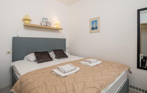 Un lit bien fait, recouvert d’une courtepointe beige, avec deux oreillers et des serviettes pliées sur un matelas blanc. Une petite étagère se trouve au-dessus du lit, sur laquelle reposent une lampe et quelques objets décoratifs. Une image encadrée est accrochée au mur, à droite du lit.