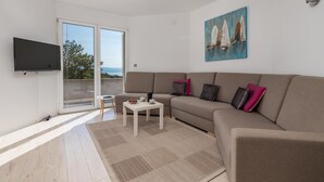 Een moderne woonkamer met een beige bank, een flatscreen televisie, een kleine tafel en een groot raam met uitzicht op zee.