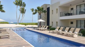 Un área junto a la piscina con sillones reclinables y sombrillas, palmeras y un edificio con balcones.