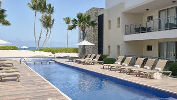 A poolside area with lounge chairs and umbrellas, palm trees, and a building with balconies.