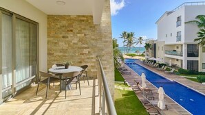 A balcony with a stone wall, a pool with lounge chairs, and a view of a building with palm trees. - Oceanfront Superior Condo with Pool in Punta Cana (Punta Cana)