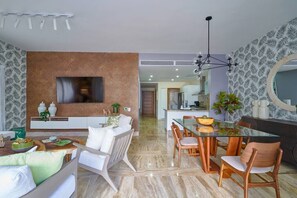 A modern living room with a TV, sofa, dining table, chairs, and a kitchen area in the background.