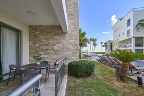 A balcony with a stone wall, a small table with chairs, a pool with lounge chairs, and a building with balconies.