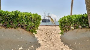 Un sendero arenoso bordeado de follaje verde que lleva a una masa de agua con un barco a lo lejos.