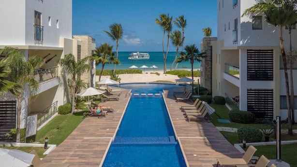 Una piscina con sillas de descanso y sombrillas, vista a la playa y palmeras.