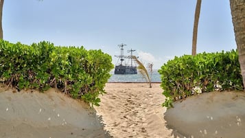 Un sentier de sable bordé de verdure mène à un plan d’eau où l’on aperçoit un navire au loin.