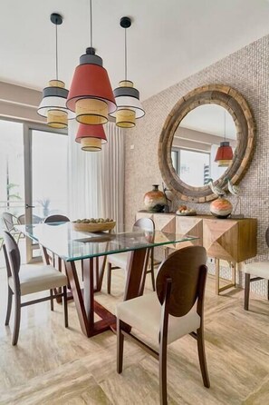 A dining area with a glass table, wooden chairs, a round mirror, and pendant lights.