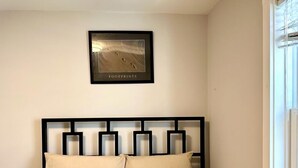 Un lit avec un cadre en métal noir et une literie beige, un tableau encadré représentant des empreintes de pas sur le mur, et une fenêtre sur la droite.