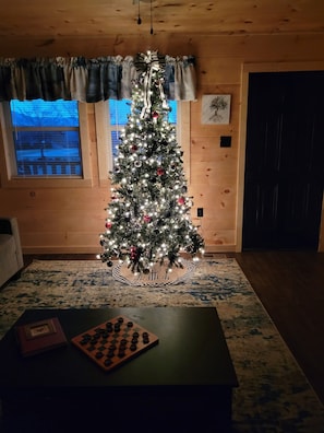 Una habitación con decoración de madera, un árbol de Navidad con luces y adornos, una ventana con persianas y un tablero de ajedrez sobre una mesa.