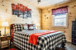 Un dormitorio acogedor estilo cabaña con colcha a cuadros, dos lámparas de noche, una manta roja y una ventana con cortina escocesa.