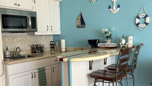 A kitchen with white cabinets, a microwave, a sink, and a bar area with two stools. There are decorative wall hangings and a ship wheel on the wall. - Beachfront condo with spectacular view (Corpus Christi)