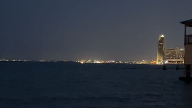 A nighttime view of a coastal area with a city skyline in the distance, a beach, and a structure with a light on top. - Beachfront condo with spectacular view (Corpus Christi)
