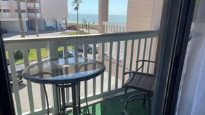 A balcony with a glass table and two chairs, overlooking a street with buildings and a palm tree. - Beachfront condo with spectacular view (Corpus Christi)