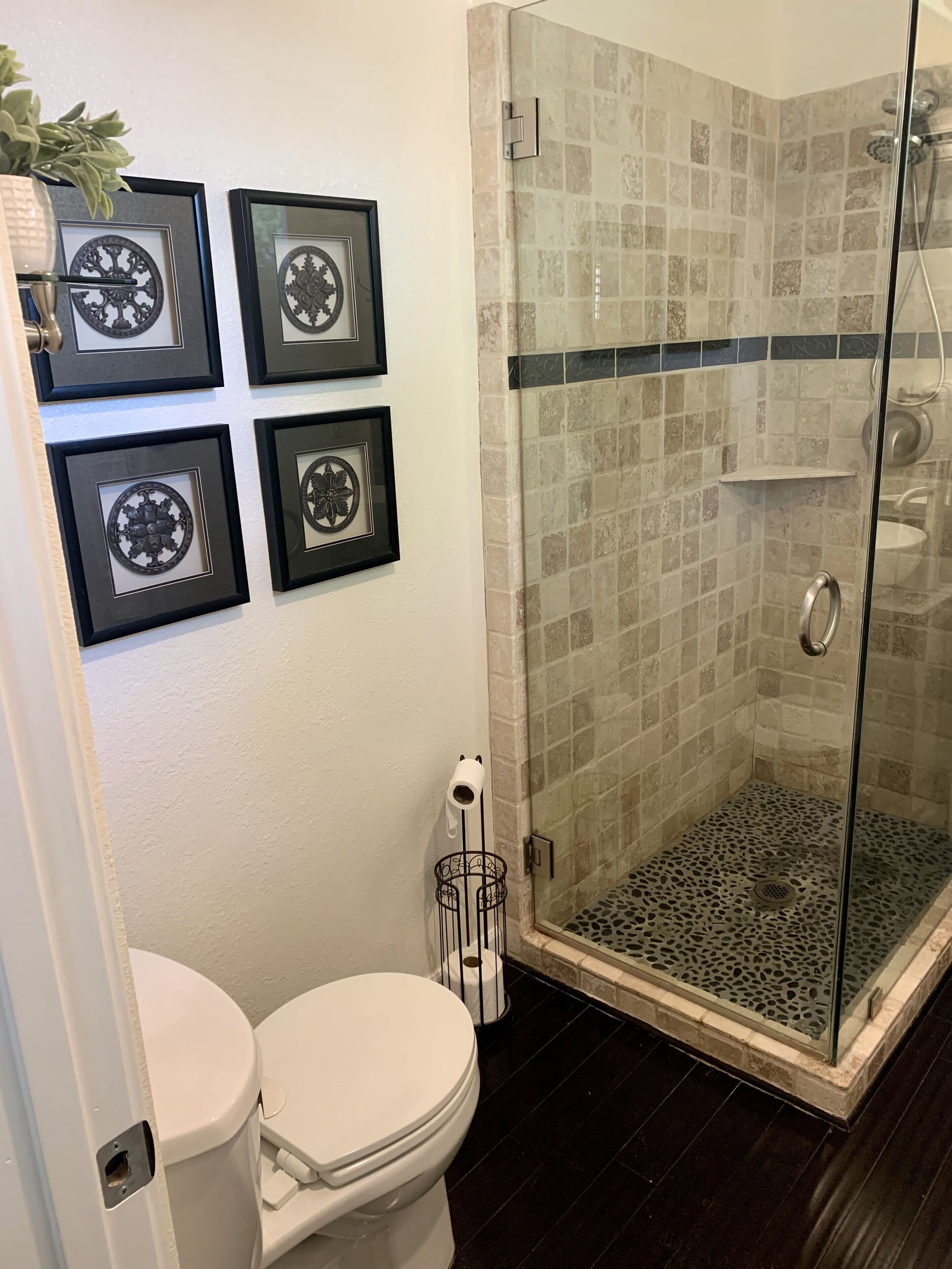 Un baño con inodoro, ducha con cerramiento de vidrio y piso de baldosas, y cuatro adornos enmarcados en la pared.